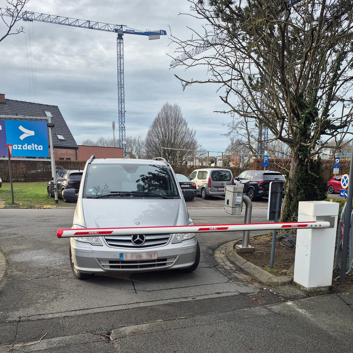 Witte Mercedes-auto geblokkeerd door rode en witte slagboom op parkeerplaats met laadpalen en bouwkraan op achtergrond.