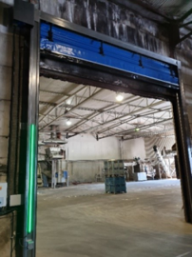 Industrieel magazijngebouw met blauwe snelrolpoort geopend, zichtbaar betonnen vloer en stellagekasten binnenin.