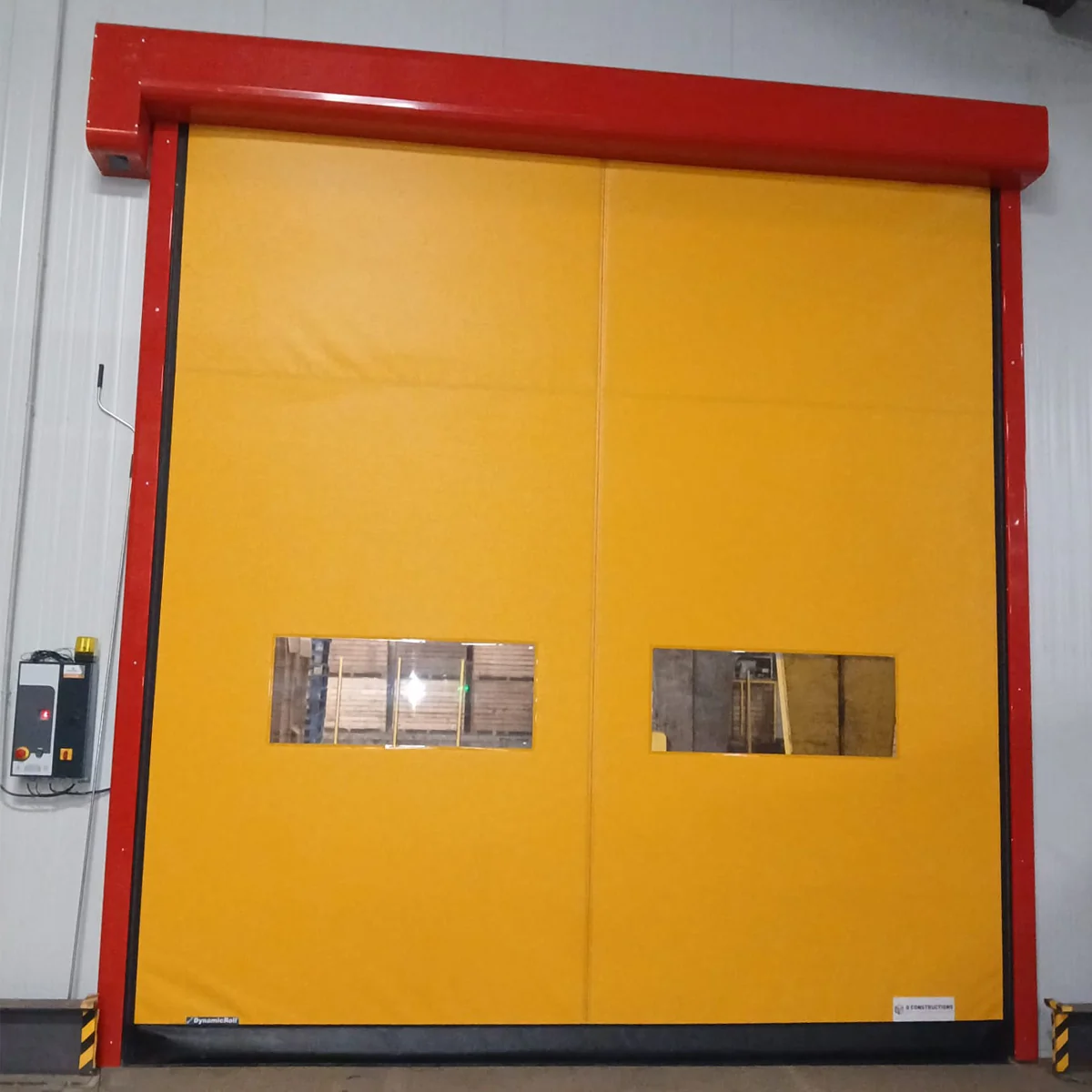 Gele snelrolpoort met rood frame en twee inspectieramen, gemonteerd in een industrieel pakhuis.