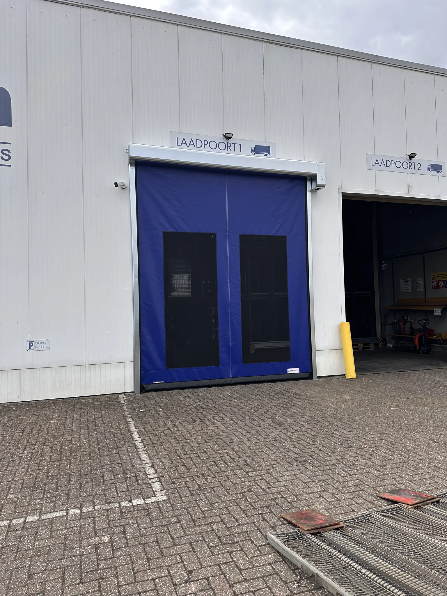 Twee laaddeuren met blauwe vliegengaaspoorten aan een industrieel gebouw, waarvan laadpoort 1 gesloten is en laadpoort 2 open.