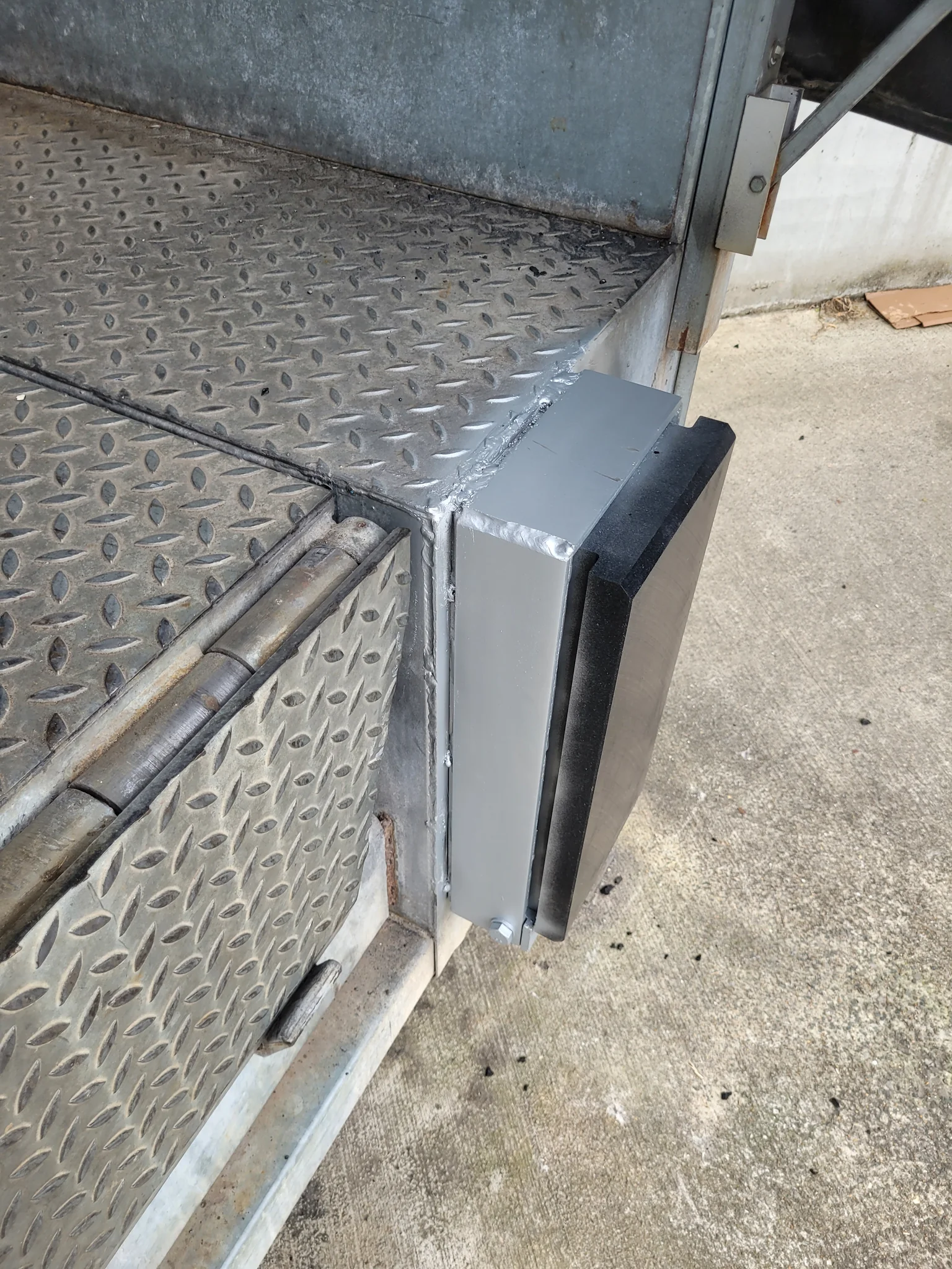 Grijs-witte stootbuffer met zwarte rubberen strip bevestigd aan de zijkant van een voertuig met geruit metalen oppervlak.