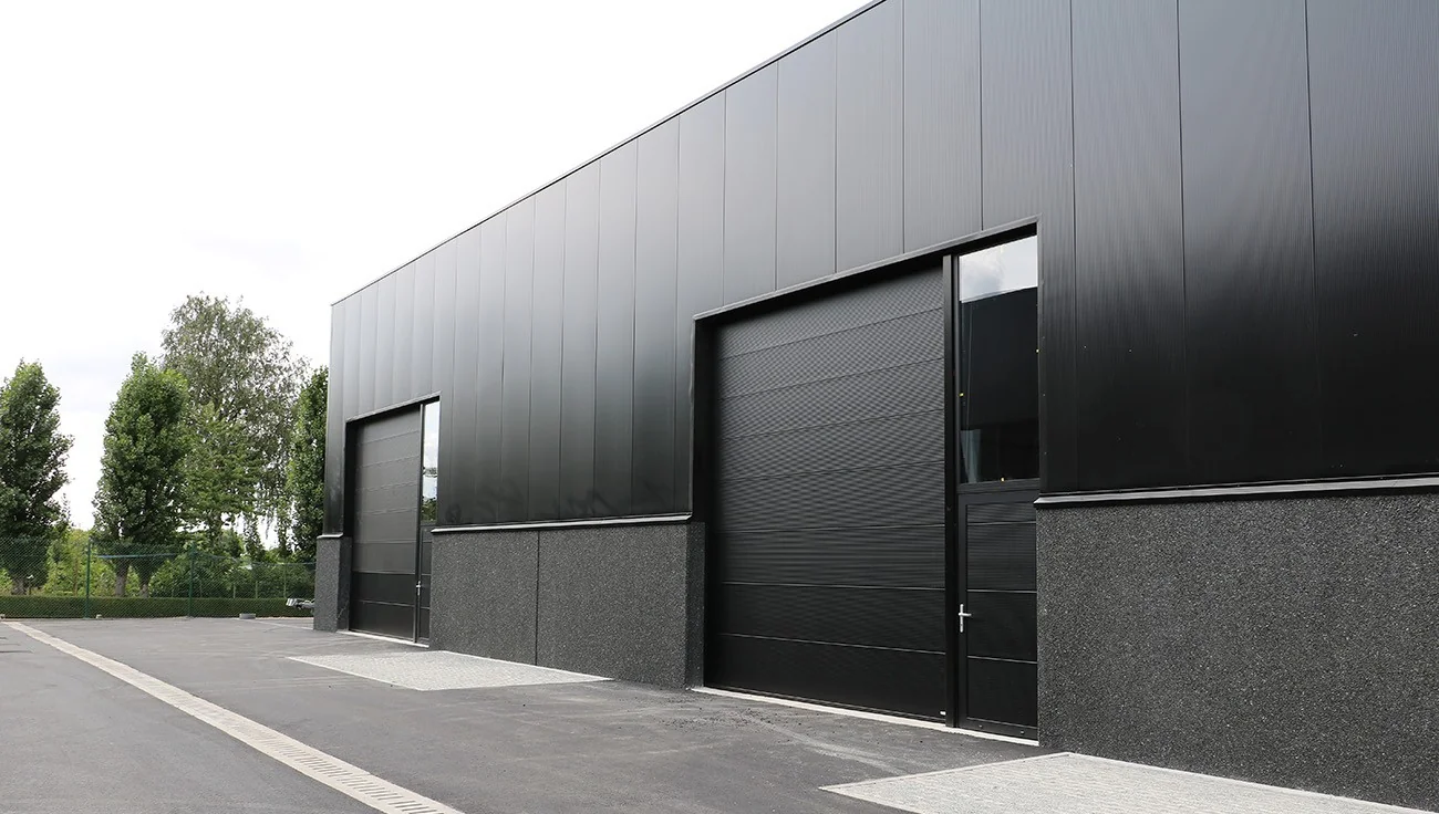 Moderne industriële garagebouw met zwarte sectionaalpoorten en grijze natuursteenvoering langs een asfaltweg.