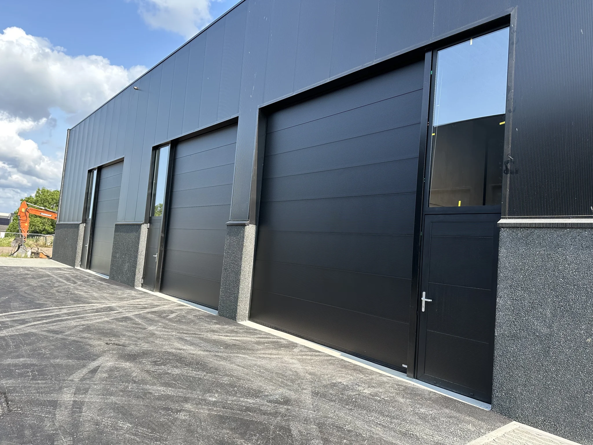 Moderne grijze industriële garagebouw met drie zwarte sectionaaldeuren en betonnen sokkel aan geasfalteerde toegang.