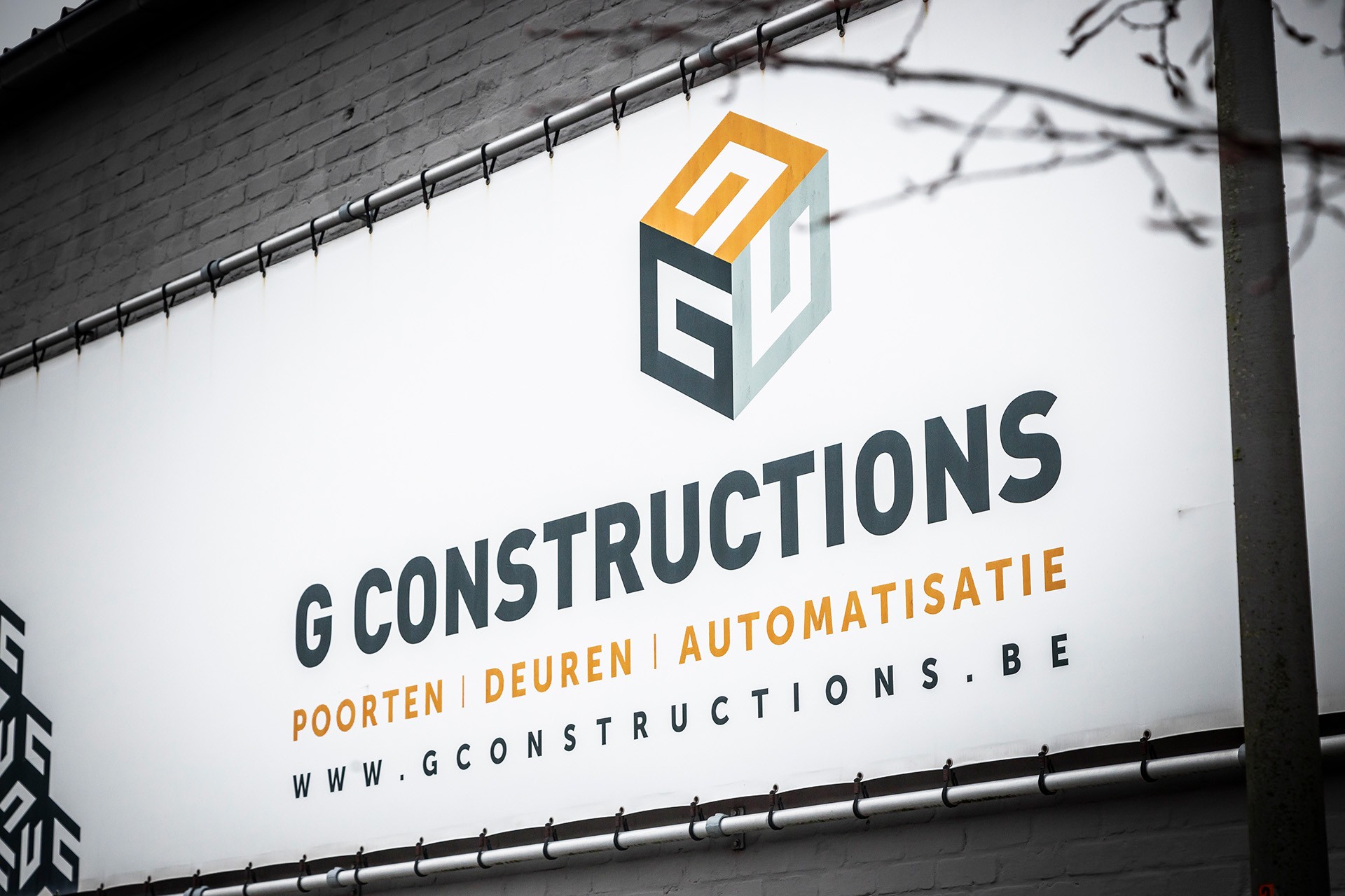 Bedrijfsbord van G Constructions met logo en tekst over poorten, deuren en automatisatie tegen bakstenen muur.