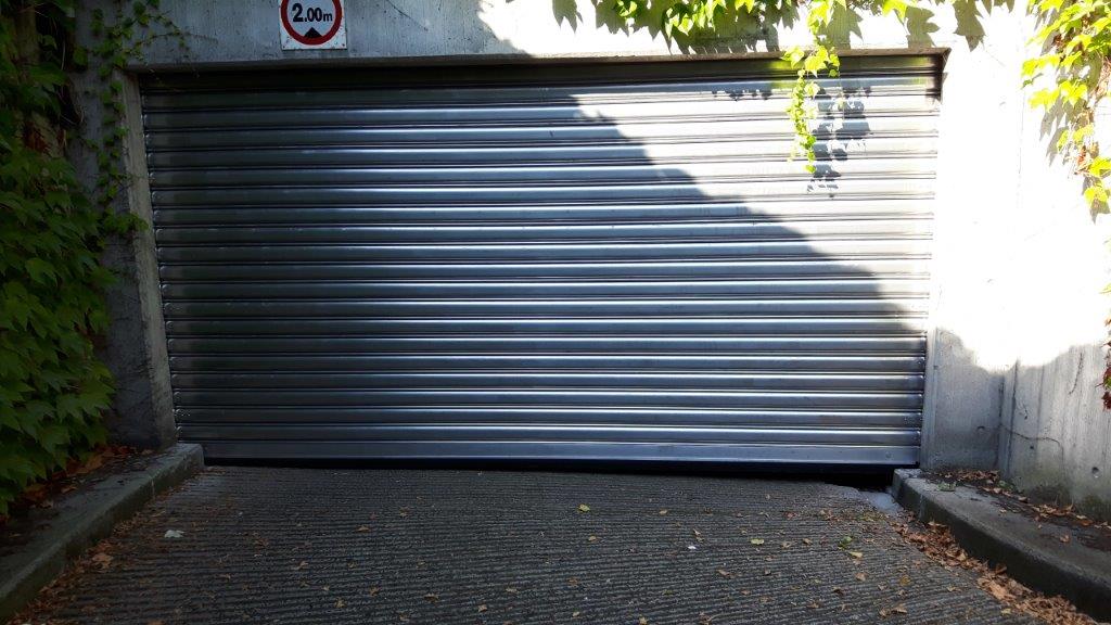 Gesloten roldeur in donkergrijs aluminium met horizontale lamellen op een oprit, omgeven door betonnen muren en klimop.
