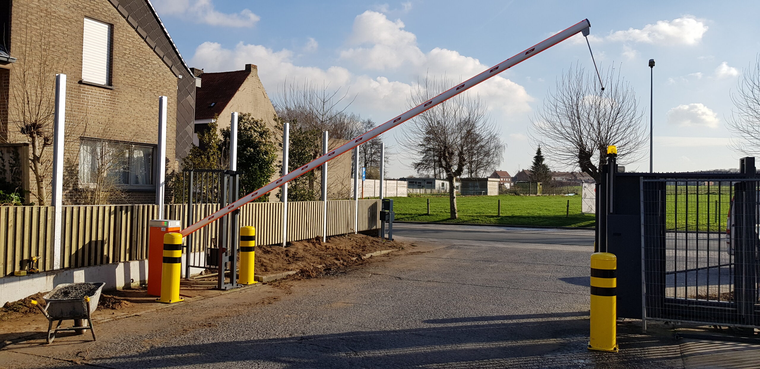 Slagboom met rood-witte arm blokkeert de ingang van een beveiligde parking naast een bakstenen huis met gele paaltjes.