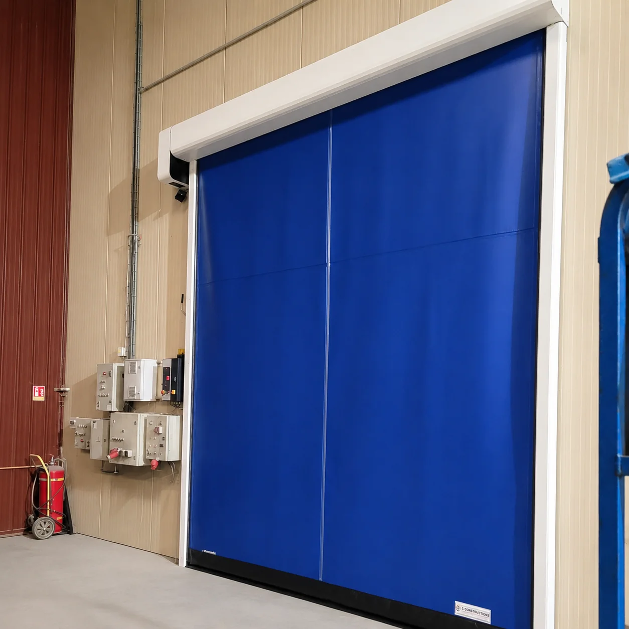 Blauwe snelrolpoort met witte frame in een industriële hal met beige golfplaten wanden en veiligheidselementen.