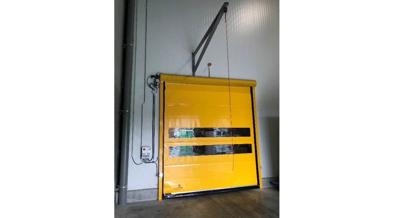 Gele snelrolpoort met zwarte horizontale strepen en transparante ramen, opgehangen aan een hijskraan in een industrieel pand.