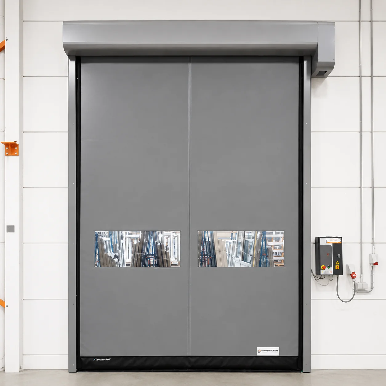 Grijze snelrolpoort met twee doorzichtige ramen in een witte industriële muur, met bedieningspaneel en veiligheidsuitrusting ernaast.