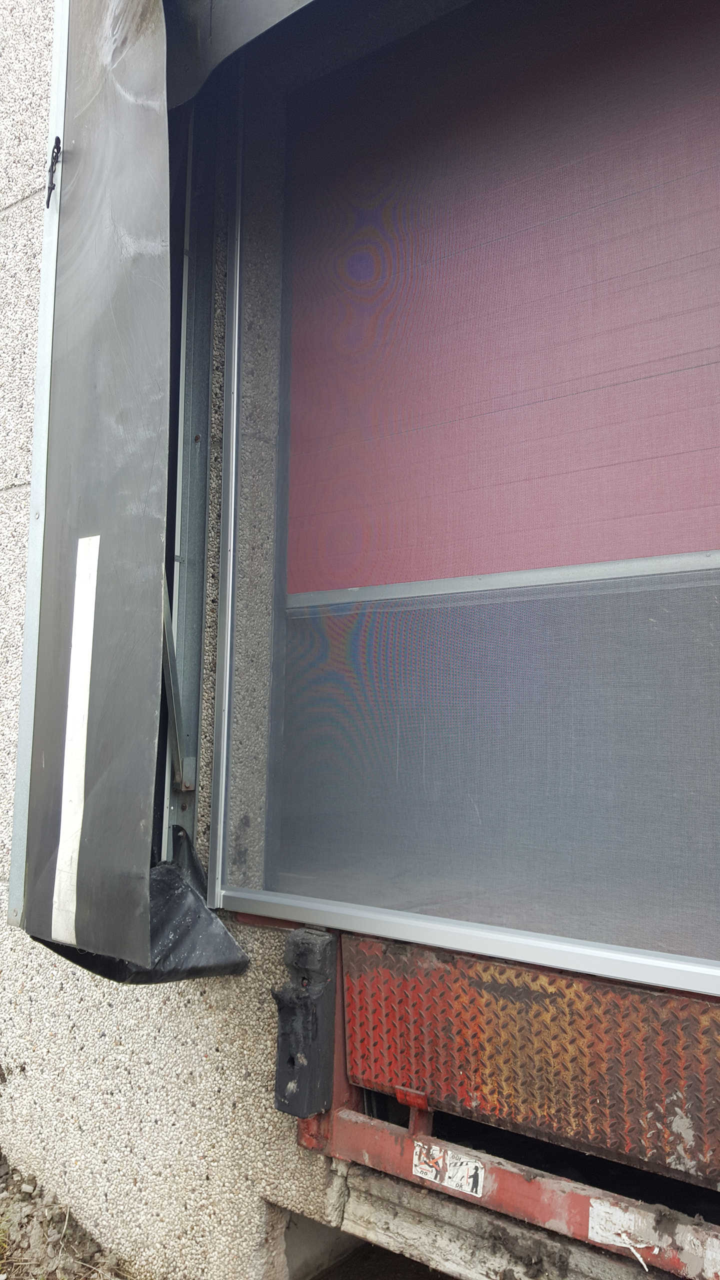 Zijkant van een rood-zwarte brandwerende poort met gaas en metalen afdekkingen tegen een betonnen muur.